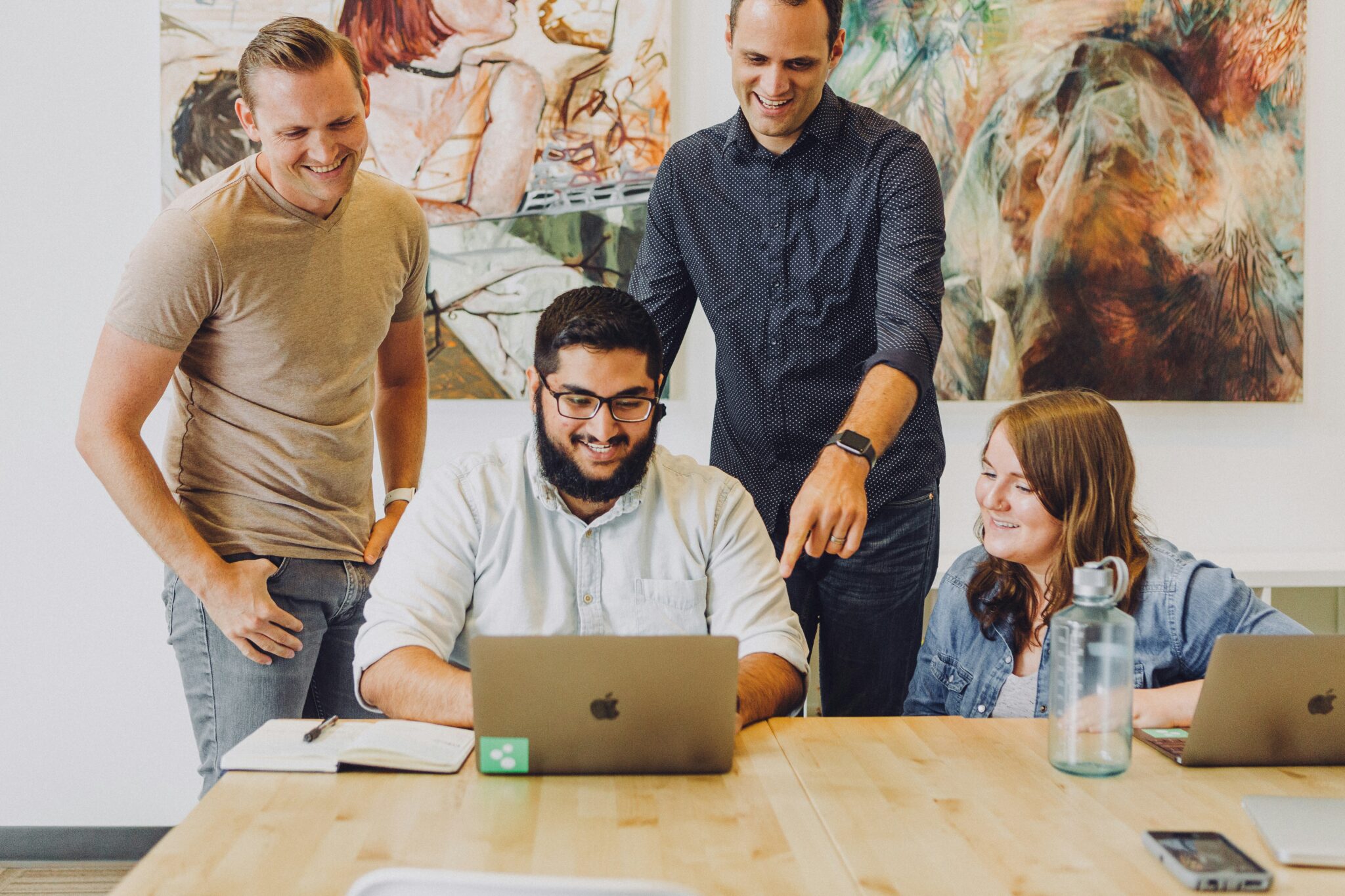Kollegen schauen auf ein MacBook, ein Kollege zeigt seine Arbeit, drei schauen ihm über die Schulter. Alle stehen in einem modernen Büro, an den Wänden hängen Gemälde, der Tisch ist aus Holz, die Stimmung ist positiv aufgeladen
