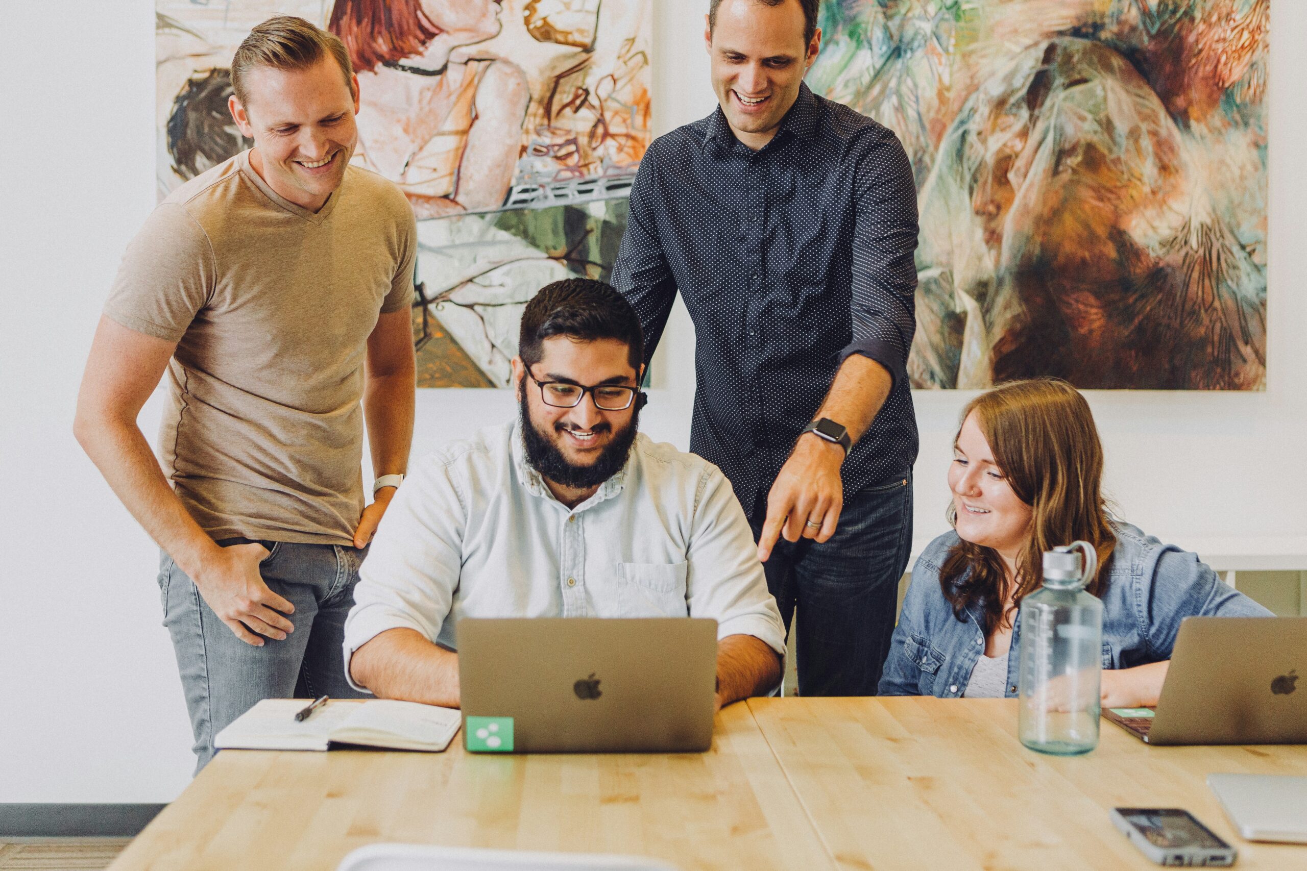 Kollegen schauen auf ein MacBook, ein Kollege zeigt seine Arbeit, drei schauen ihm über die Schulter. Alle stehen in einem modernen Büro, an den Wänden hängen Gemälde, der Tisch ist aus Holz, die Stimmung ist positiv aufgeladen